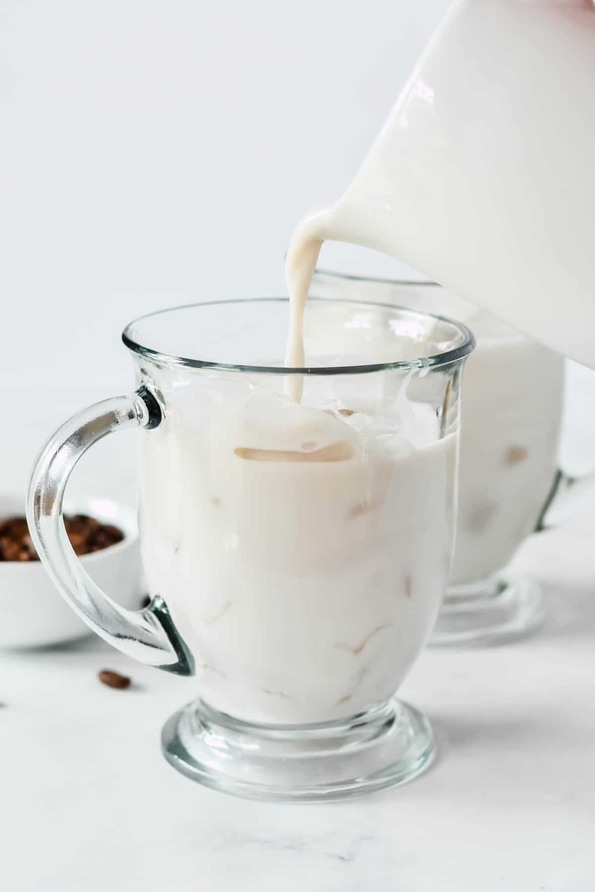 Dairy-free milk is being poured from a pitcher into a clear glass mug filled with ice.