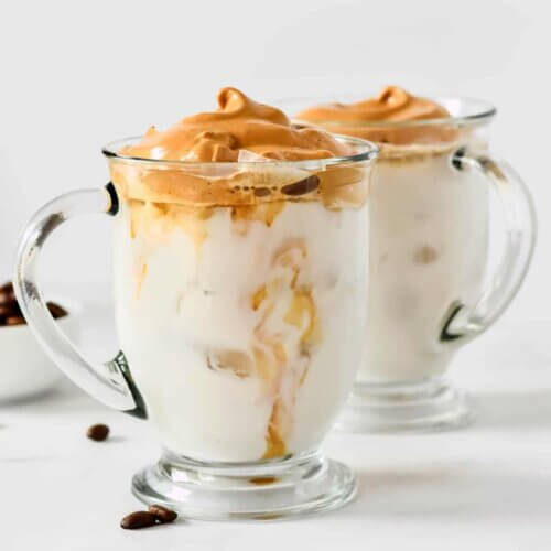 Two clear glass mugs are filled with iced milk and dolloped with thick whipped coffee.
