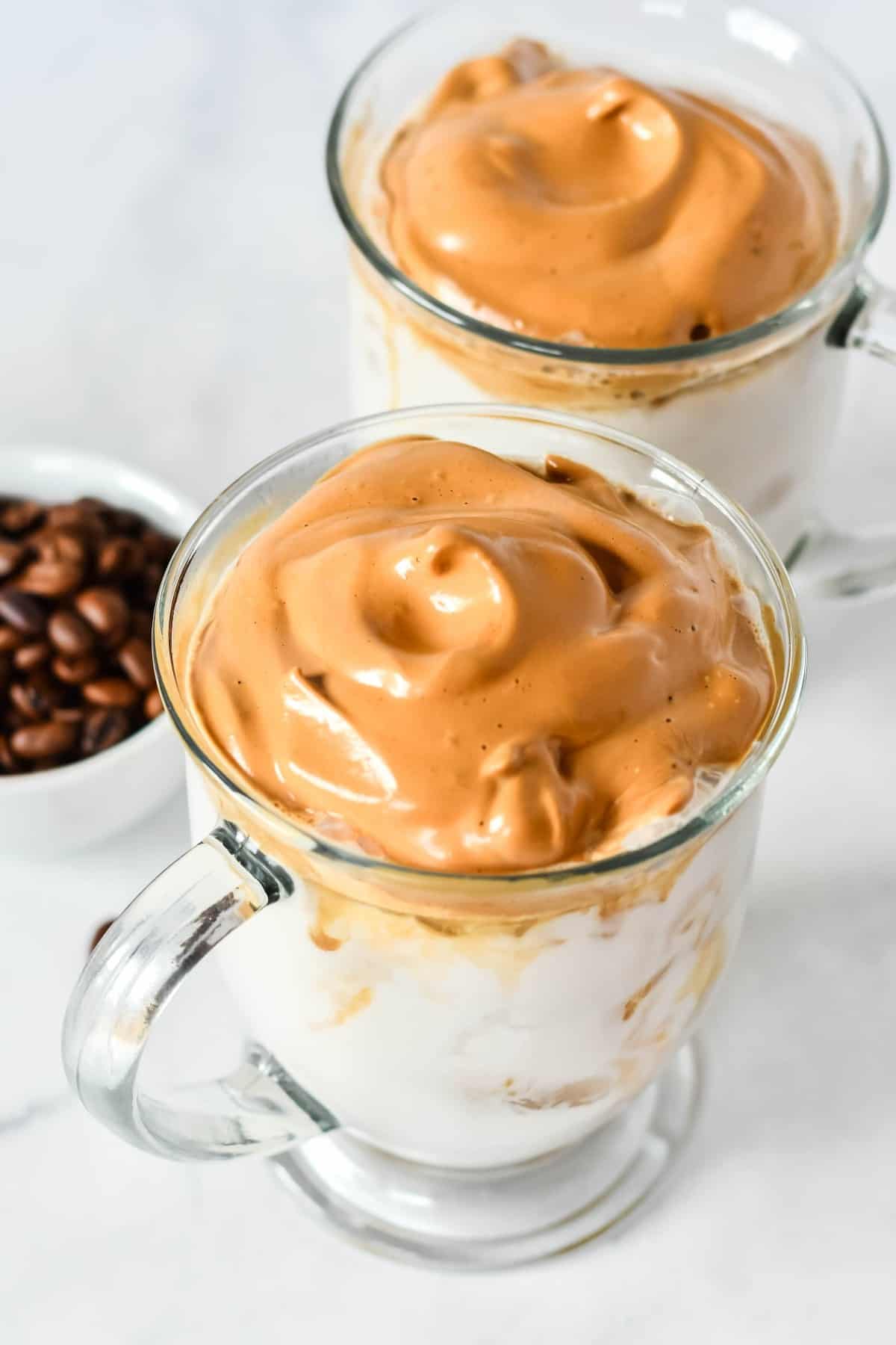Two clear glass mugs are tilted forward showing a closeup of a thick caramel colored creamy topping.