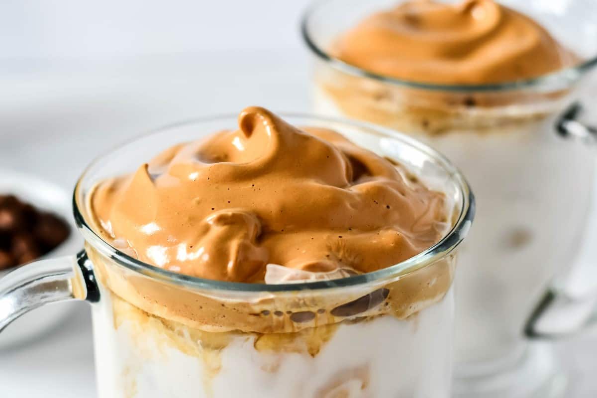 Wide photo of whipped topping on the top of iced milk in two clear glass mugs.