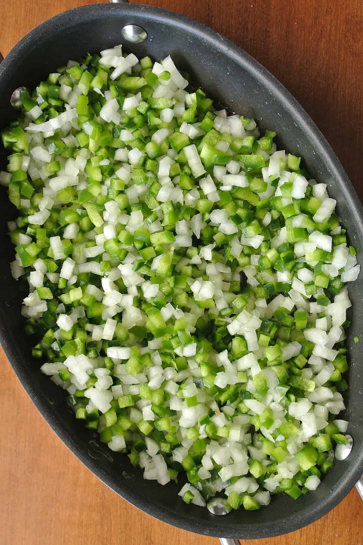Overhead view of small diced white onions and green bell peppers are in an oblong frying pan and just starting to saute.