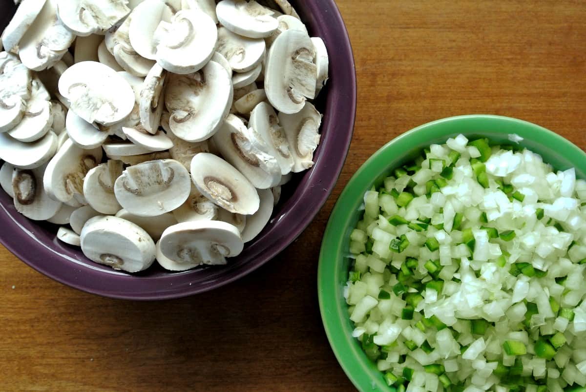 Overhead view of sliced mushrooms and diced onion and green bell peppers in their own bowls.