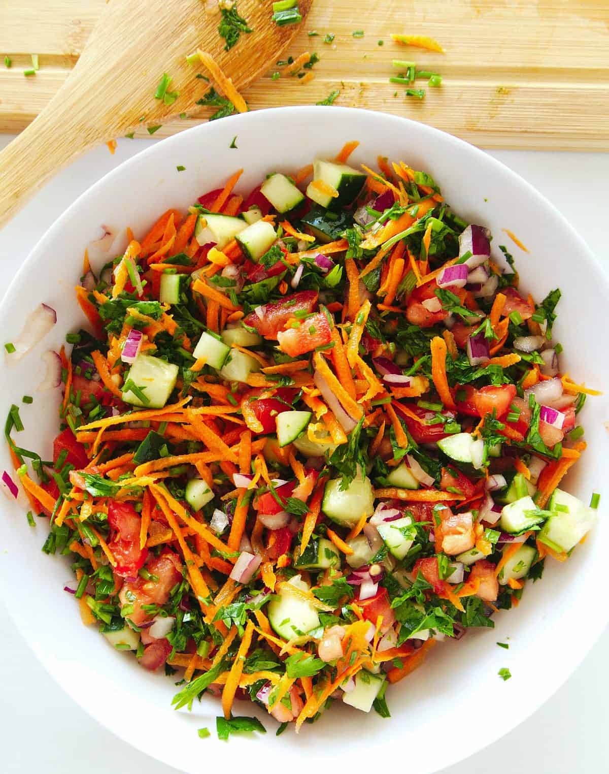 Overhead view of a cold mixed side dish in a large white bowl. On the side is a wooden spoon with salad remnants around.