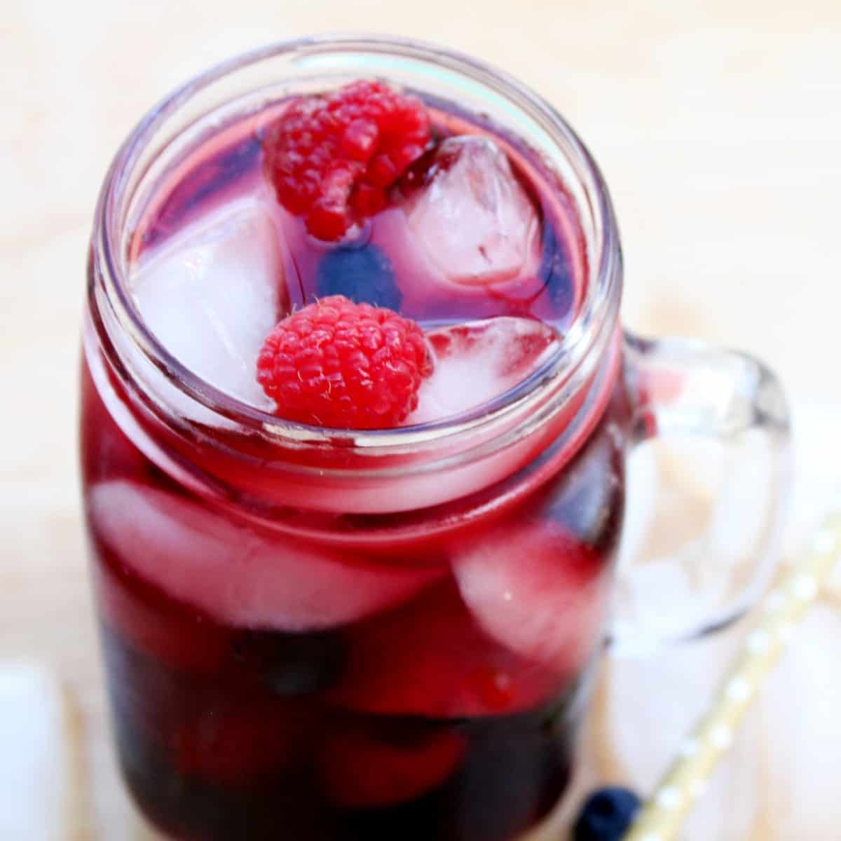 A tilted jar mug is tilted forward with red wine sangria over ice.Raspberries and blueberries are interspersed throughout.