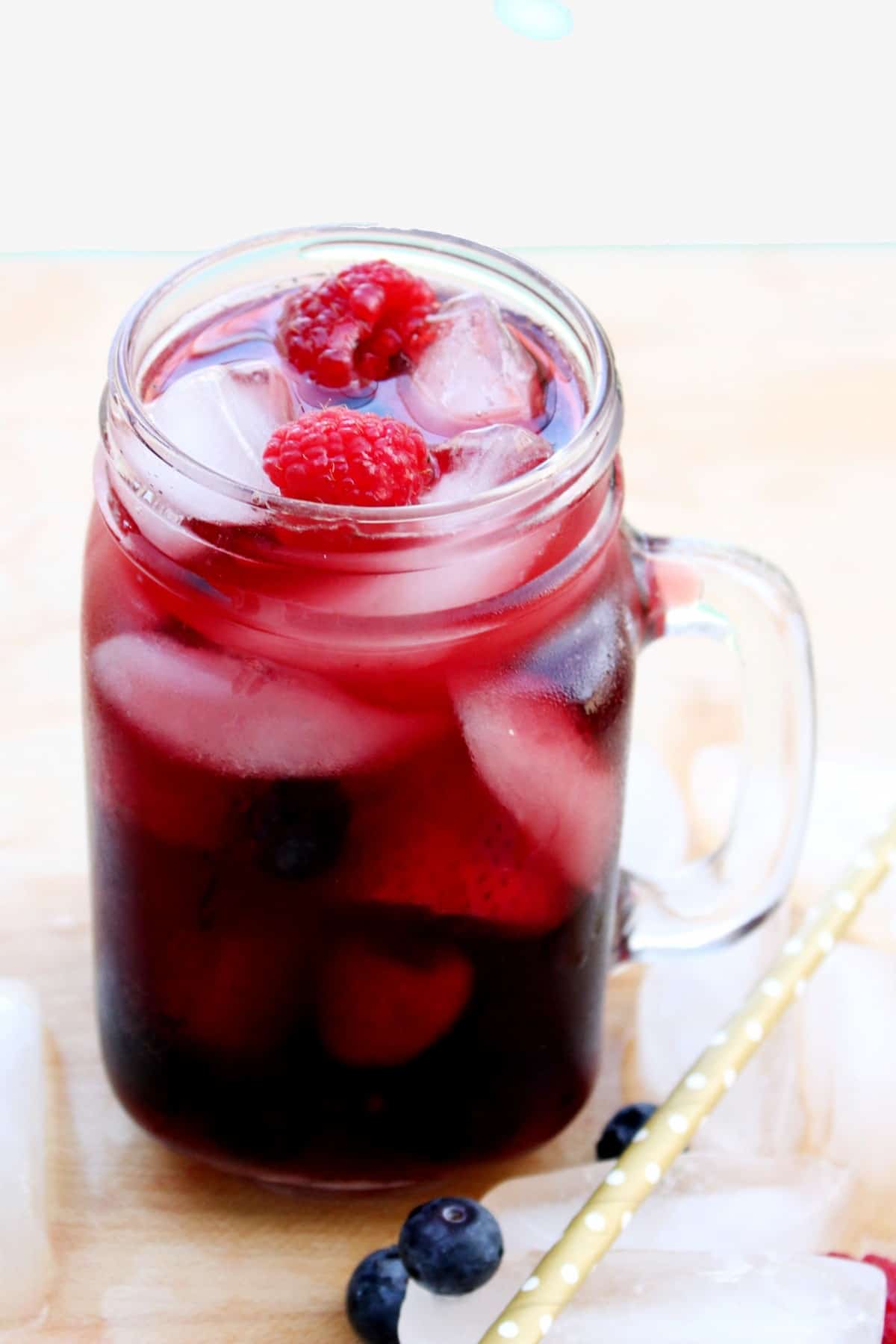 A mug glass is full of rich looking red wine with berries and ice. A straw and ice are on the table next to it.