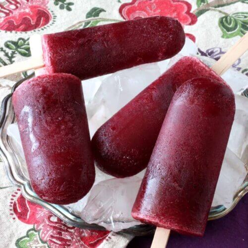 Four deep blackberry colored popsicles laying criss-cross on top of each other and on a bed of ice in a silverplate tray.