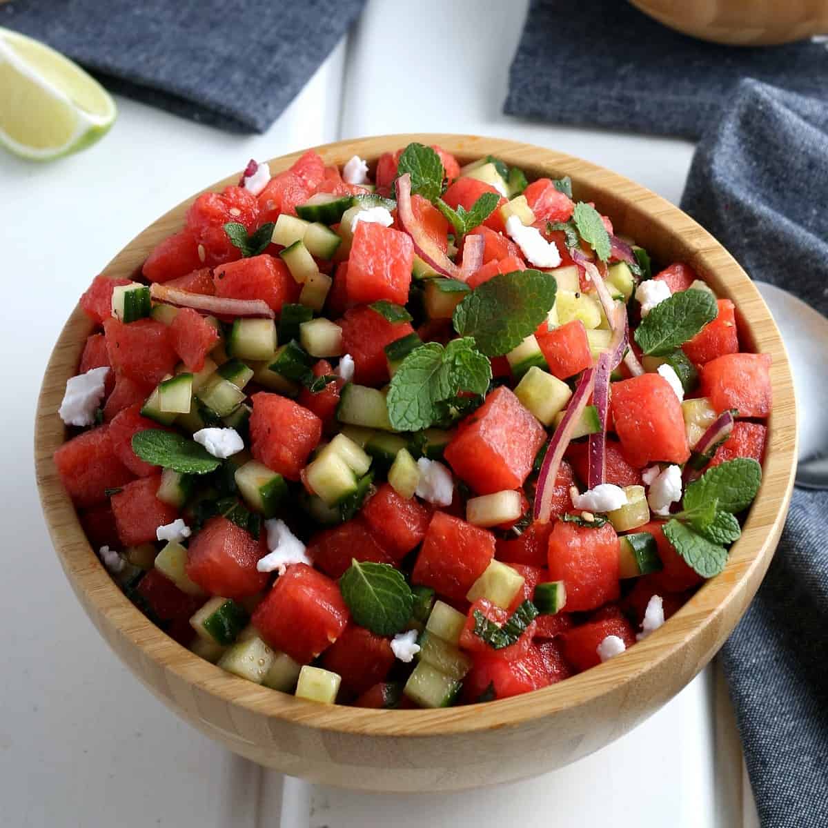 A centered wood bowl that is full of watermelon feta salad with a gray cloth napkin on the side.
