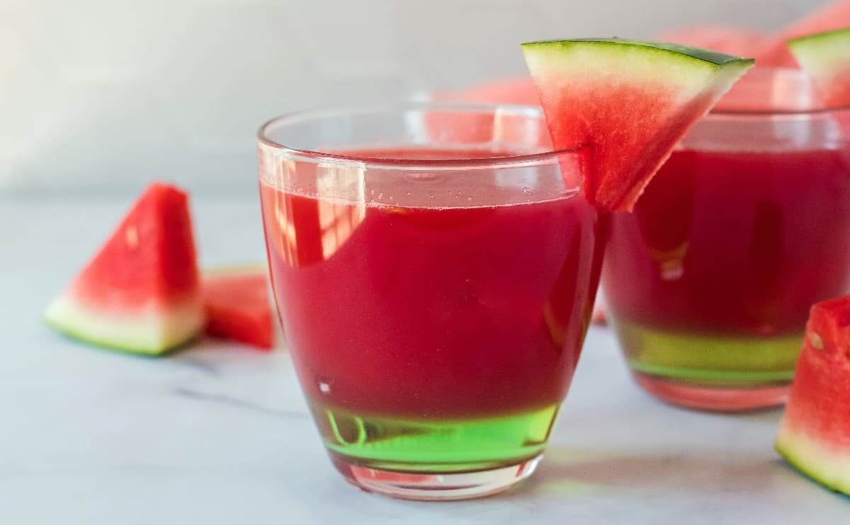 Wide photo showing two glasses filled with a red cocktail and extra watermelon wedges are laying around.