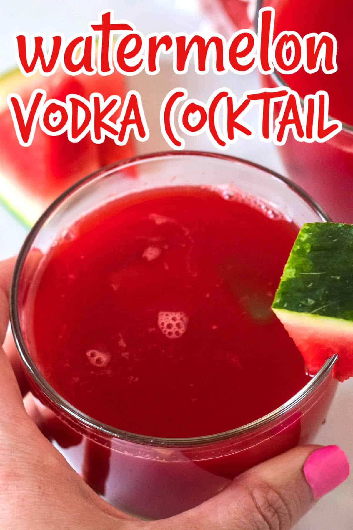 A hand is holding a watermelon red cocktail very close to the camera lens with a watermelon wedge as garnish.
