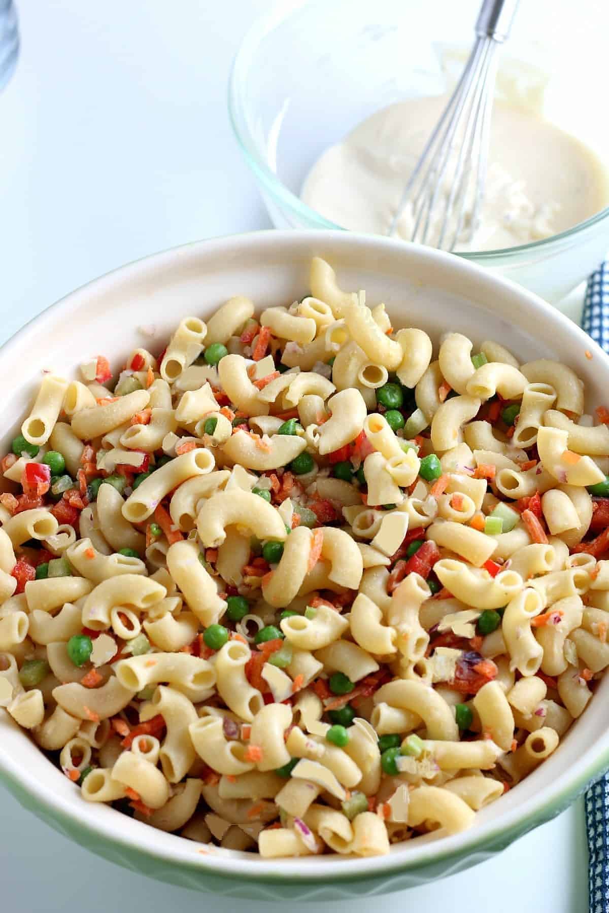 A large bowl full of old fashioned macaroni salad and small chopped veggies waiting to be tossed with the creamy dressing in a bowl that's in the background.