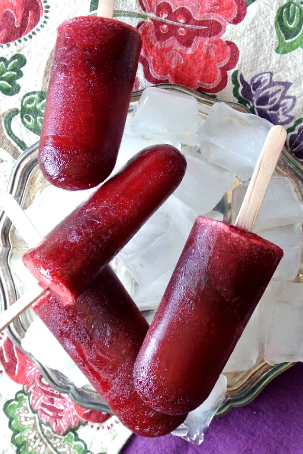 Four deep blackberry colored popsicles laying criss-cross on top of each other and on a bed of ice in a silverplate tray on on a brightly colored flowered cloth napkin.