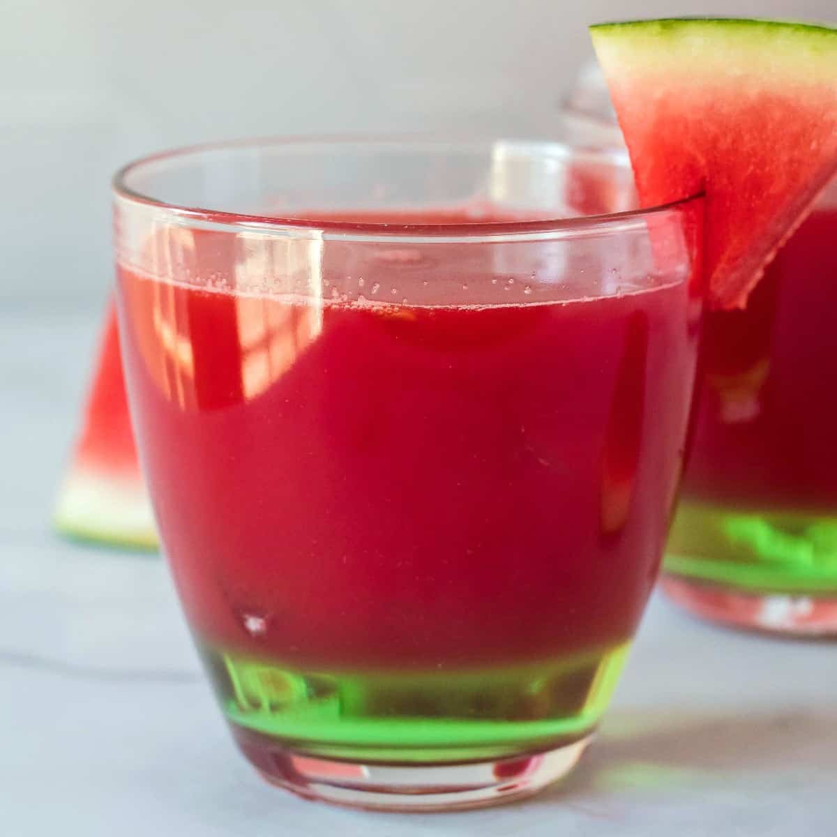 A bright red drink is filling a short tumbler and has mini fruit garnish around.