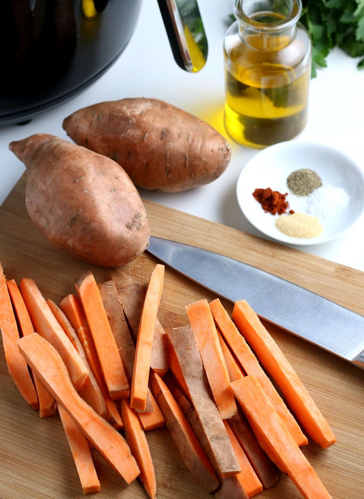 Sliced sweet potatoes in a french fry shape on a wooden board with a knife.