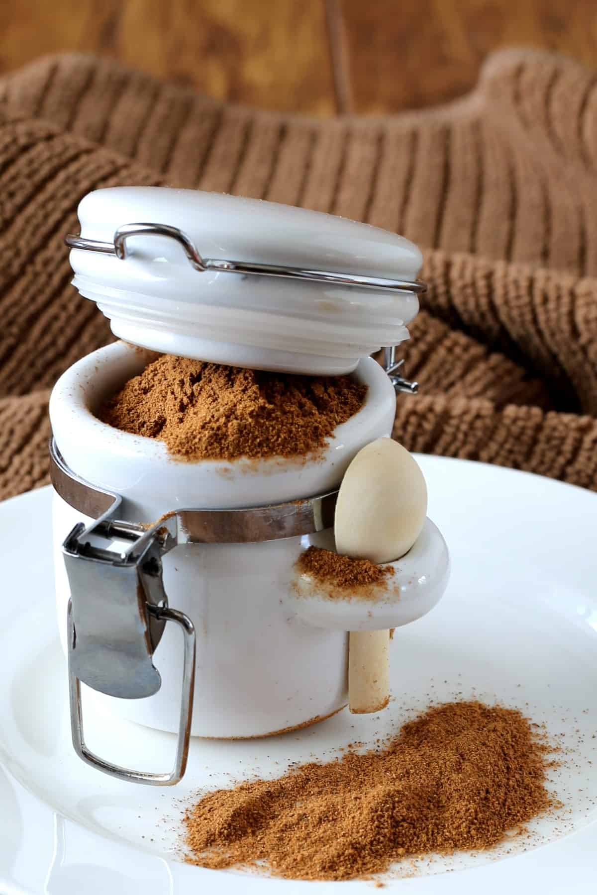 Small white jar with spoon in slot and spices overflowing onto white plate.