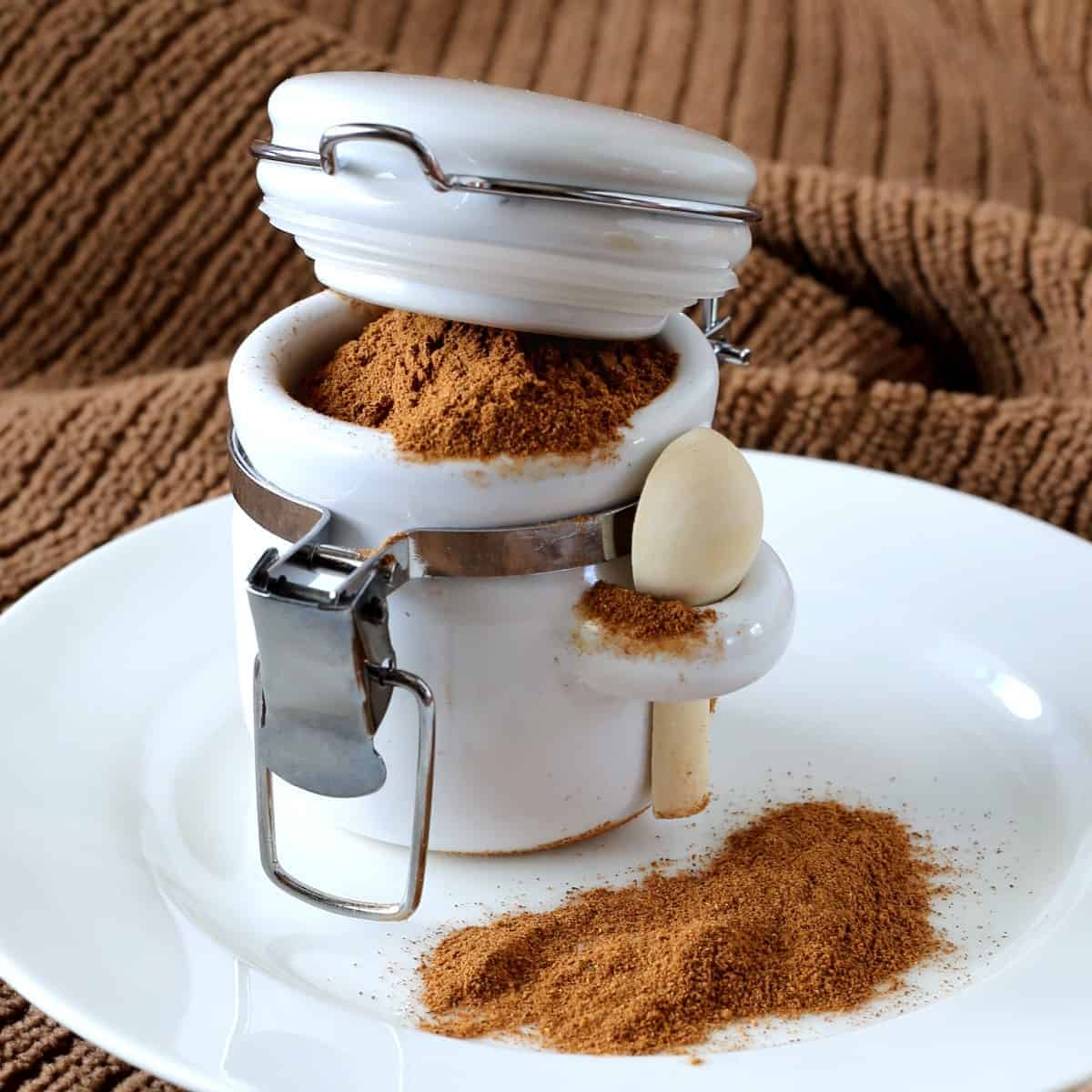 Small white jar with a hooked spoon and pumpkin seasoning blend is overflowing.