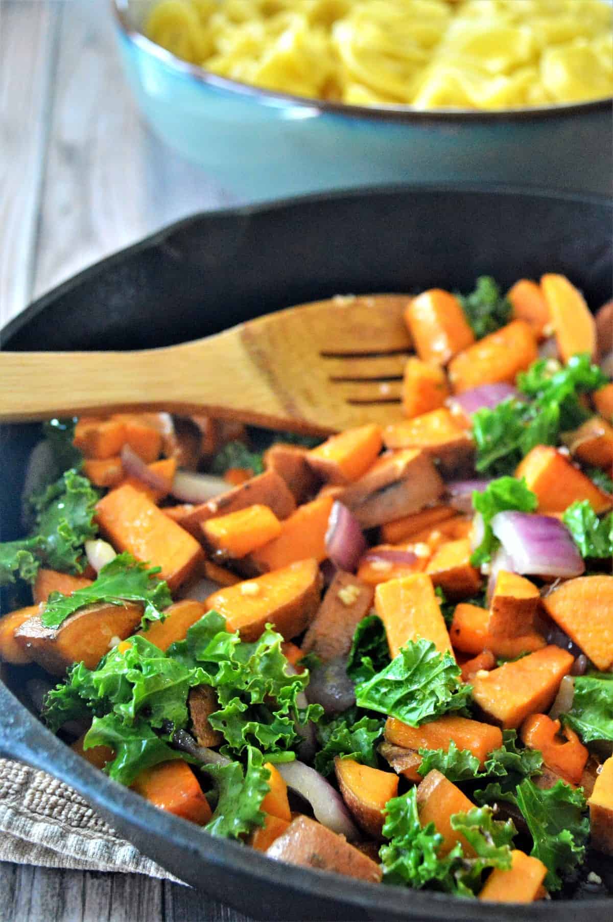 Skillet of vegetables and kale being cooked and stirred with a wooden spoon.