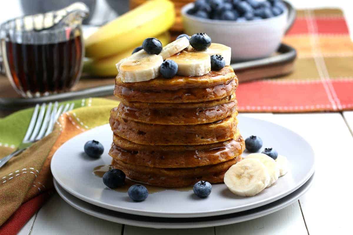 Stack of waffles in front of syrup, bananas, and blueberries.