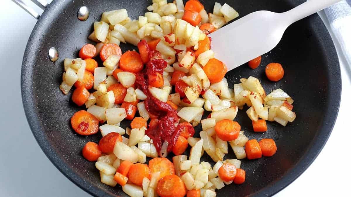 Skillet full of browned carrot and potatoes chunks with tomato paste being added.