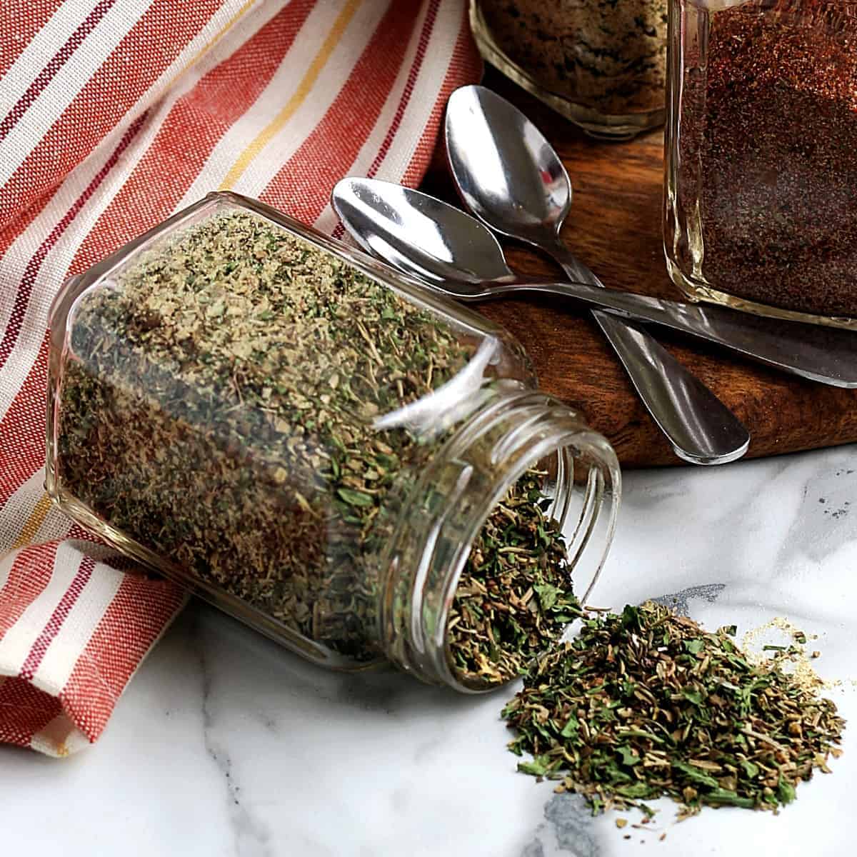 Jar of spilled homemade Italian seasoning in front of two jars of different mixes.