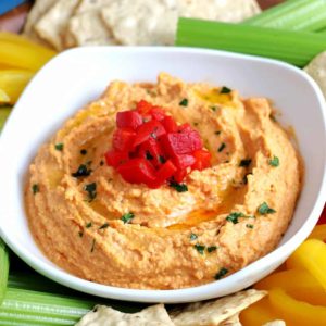Tilted white bowl full of red pepper hummus with veggies around.