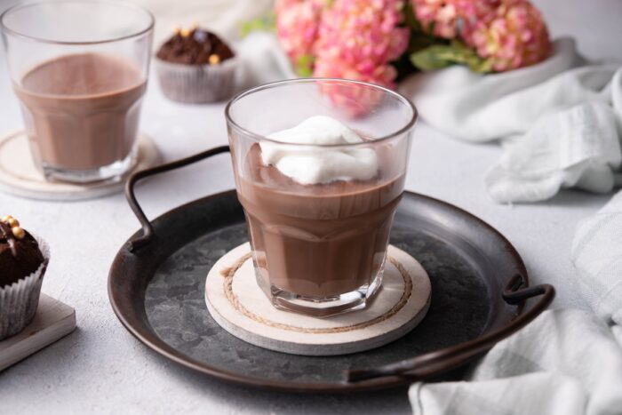 cardamom spiced vegan hot chocolate is served in a clear mug, showcasing its rich brown coloring and topping of whipped coconut cream.